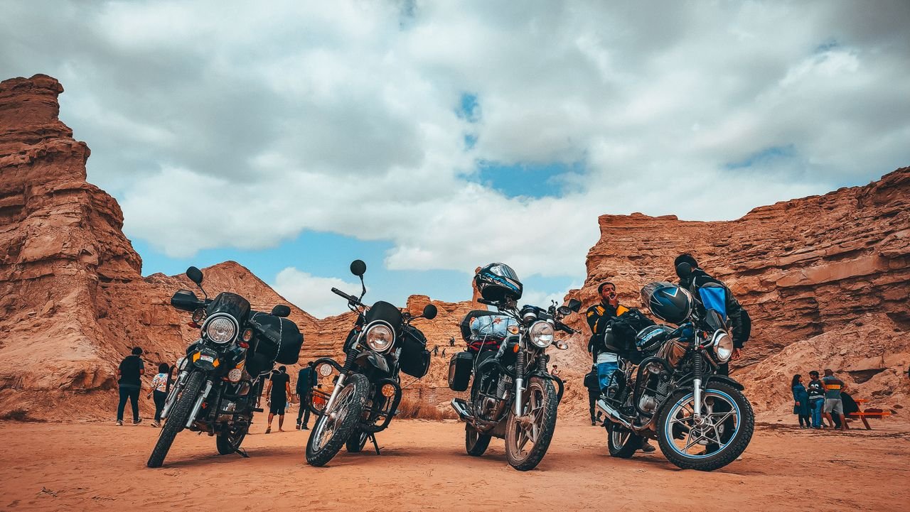 Home A group of parked motorcycles and riders in a scenic desert canyon under a cloudy sky.